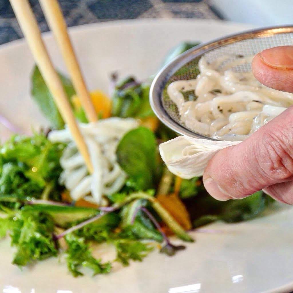 Chopsticks lifting noodles over a strainer, beside a fresh salad